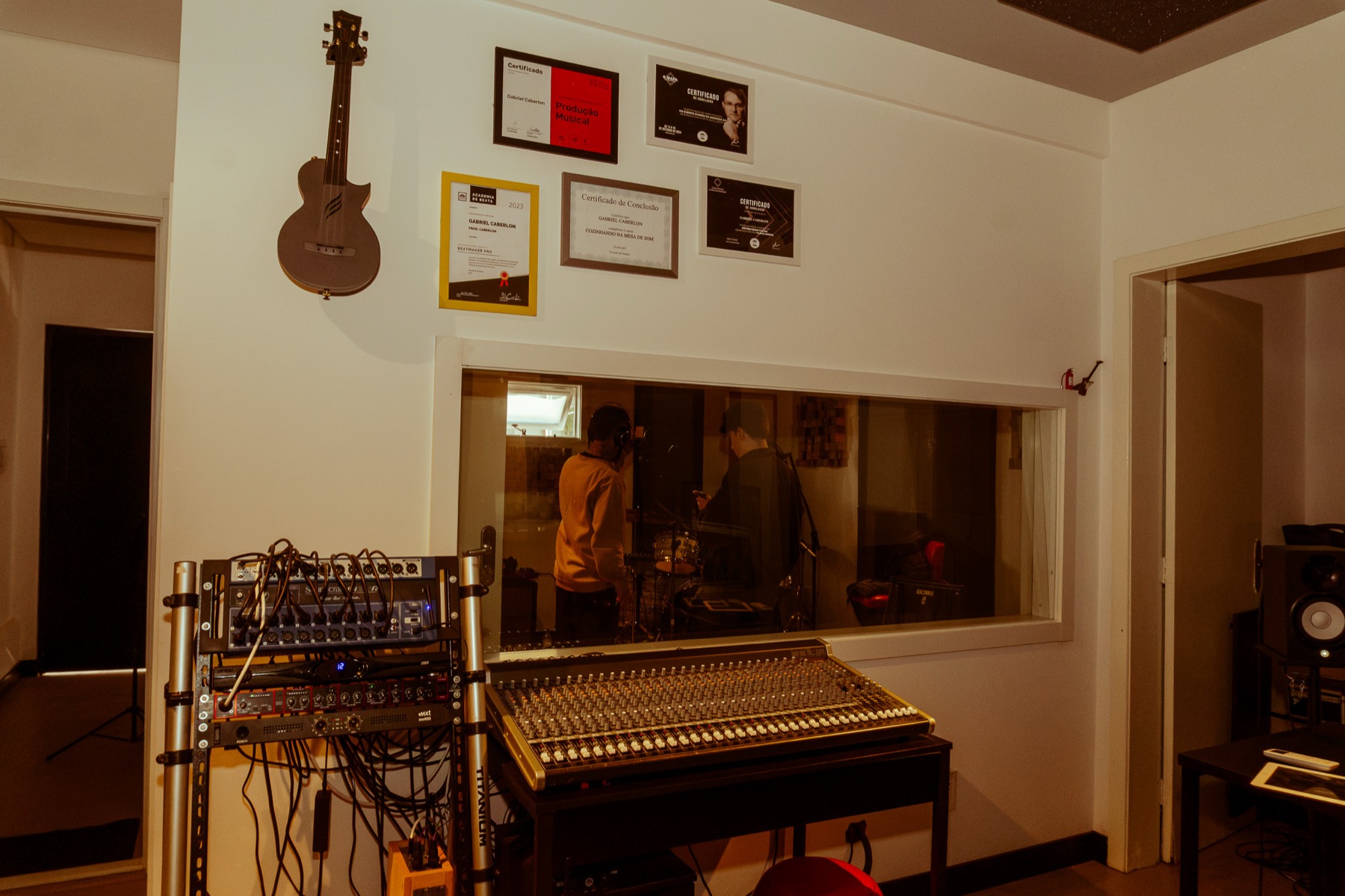 Mesa de som em primeiro plano e músicos gravando bateria na sala ao fundo, vistos pelo visor